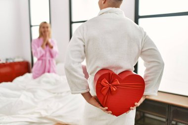 Sevgilisine sürpriz yapan bir adam sevgilisinin hediye kutusuyla evde yatağında oturuyor..