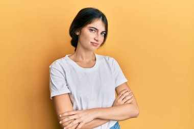Gündelik beyaz tişört giyen genç beyaz kadın şüpheci ve gergin, yüzünde çapraz kollu tasvip etmeyen ifade. negatif kişi. 