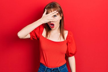 Kırmızı tişörtlü, kızıl saçlı genç bir kadın. Şoka girmiş, yüzünü ve gözlerini kapamış, utanmış bir ifadeyle parmaklarından bakıyor.. 