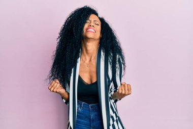 Orta yaşlı Afro-Amerikalı kadın gündelik kıyafetler giyip çok mutlu ve heyecanlı bir şekilde kollarını kaldırıp, gülümseyerek ve başarı için bağırarak kazanan jestler yapıyor. kutlama konsepti. 