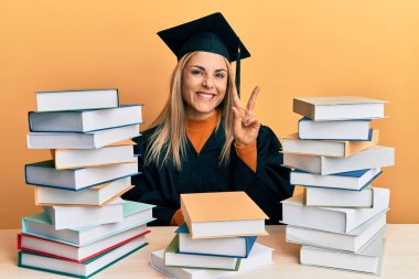 Mezuniyet cüppesi giyen genç beyaz kadın masanın üzerinde oturuyor ve parmaklarını iki numara ile göstererek gülümsüyor. Kendinden emin ve mutlu.. 