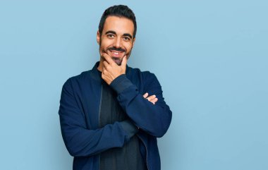 Gündelik kıyafetler giyen genç İspanyol adam kameraya kendinden emin bakıyor kollarını kavuşturup gülümsüyor ve çenesini kaldırıyor. Olumlu düşünmek. 