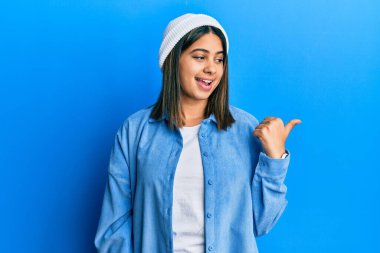 Şirin yün şapkalı, mutlu yüzlü, gülümseyen ve başparmağıyla yan tarafı gösteren genç Latin kadın.. 