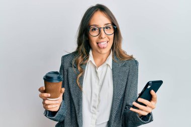 Genç esmer kadın akıllı telefon kullanıyor ve bir fincan kahve içiyor. Dilini komik bir ifadeyle dışarı çıkarıyor.. 