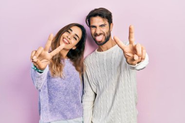 Latin kökenli genç bir çift, günlük kıyafetler giyip gülümsüyor ve her iki elin parmaklarını zafer işareti yaparken gösteriyor. İki numara.. 