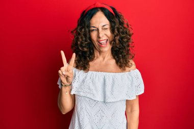 Orta yaşlı İspanyol kadın gündelik kıyafetler giyip kameraya gülümseyen ve zafer işareti yapan. İki numara.. 