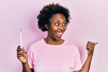 Genç Afro-Amerikalı kadın elinde elektrikli diş fırçası, baş parmağını yana uzatıyor, ağzı açık gülümsüyor. 