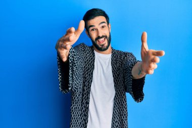 Gündelik giysiler giymiş genç İspanyol adam kameraya bakıp sarılmak için kollarını açmış gülümsüyor. Mutluluğu kucaklayan neşeli bir ifade.. 