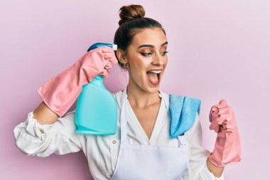 Önlük giymiş güzel esmer genç bir kadın temizlik spreyi tutarak baş parmağıyla yan tarafa işaret ederek ağzı açık gülümsüyor. 