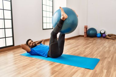Yakışıklı İspanyol adam egzersiz yapıyor ve yoga minderinde geriniyor, spor salonunda pilates topuyla esneklik gösteriyor.