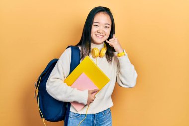 Öğrenci çantası ve kitapları tutan genç Çinli kız telefonla el ve parmak hareketleriyle gülümsüyor telefonda konuşur gibi. iletişim kavramları. 