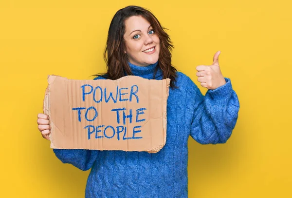 Young plus size woman holding power to the people banner smiling happy and positive, thumb up doing excellent and approval sign