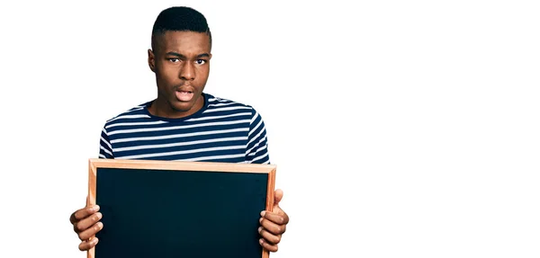 衝撃的な顔で黒板を持っている若いアフリカ系アメリカ人の男性 懐疑的で皮肉を探して 口を開けて驚いて — ストック写真