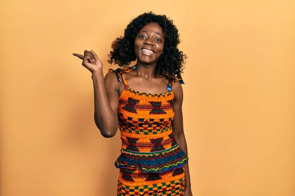 顔に大きな笑顔で伝統的なアフリカの服を着ている若いアフリカ系アメリカ人の女性 カメラを見て側に手と指を指して — ストック写真