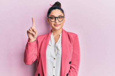 Ortadoğulu güzel bir kadın iş ceketi ve gözlük takıyor. Bir numaralı parmağını gösteriyor ve işaret ediyor. Kendinden emin ve mutlu bir şekilde gülümsüyor.. 