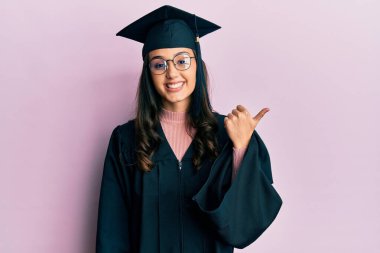 Mezuniyet şapkası ve tören cübbesi takan genç İspanyol kadın gülümseyen yüzüyle yan tarafa bakıyor ve başparmağıyla işaret ediyor.. 