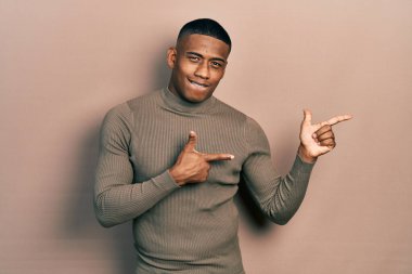 Balıkçı yaka kazak giyen genç siyahi bir adam gülümseyip kameraya bakıyor ve iki eliyle ve parmaklarıyla kenarı işaret ediyor.. 