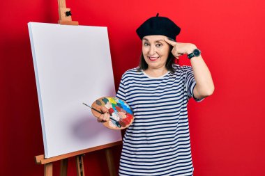 Orta yaşlı İspanyol kadın paletle resim çiziyor, resim sehpası ayakta gülümsüyor, tek parmağıyla kafasını işaret ediyor, harika bir fikir ya da düşünce, iyi bir hafıza 