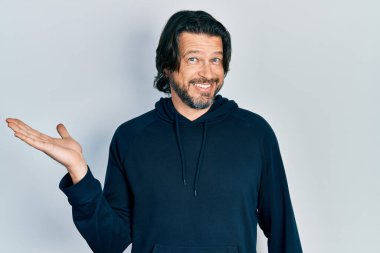 Orta yaşlı beyaz bir adam sıradan bir kazak giyiyor neşeli bir şekilde gülümsüyor ve avuç içi kameraya bakıyor.. 