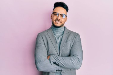 İş kıyafetleri giymiş genç bir Arap, kollarını kavuşturup kameraya bakarken gülümsüyor. pozitif kişi. 