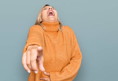 Orta yaşlı beyaz kadın, sıradan bir kazak giyip sana gülüyor, kamerayı işaret ediyor, yüzünü gösteriyor, utanç ifadesi. 