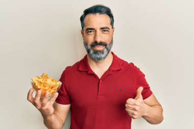 Orta yaşlı, sakallı, elinde patates cipsi tutan mutlu ve pozitif gülümseyen, başparmağını kaldıran ve onay işareti yapan bir adam. 