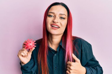 Virüs oyuncağı taşıyan genç beyaz kadın mutlu ve pozitif gülümsüyor, başparmağıyla mükemmel ve onay işareti yapıyor. 