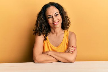 Masada oturan, kollarını kavuşturup kameraya bakan ve gülümseyen orta yaşlı güzel bir kadın. pozitif kişi. 