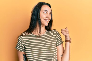 Günlük çizgili tişört giyen genç İspanyol kadın gülümseyen yüz ifadesiyle ve başparmağıyla yan tarafa bakıyor.. 