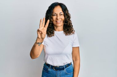 Orta yaşlı, beyaz tişört giyen İspanyol bir kadın üç numarayı gösterip işaret ederken kendinden emin ve mutlu bir şekilde gülümsüyor.. 