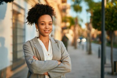 Kollarını kavuşturmuş genç Afrikalı Amerikalı iş kadını şehirde mutlu bir şekilde gülümsüyor..