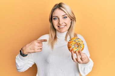 Genç beyaz kadın donut tutuyor gülümsüyor mutlu bir şekilde eli ve parmağı ile işaret ediyor 