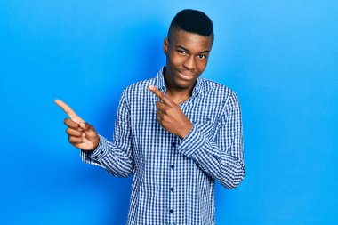 Genç Afro-Amerikalı sıradan kıyafetler giyen, gülümseyen ve kameraya bakan iki elini ve parmağını yana doğru gösteren bir adam.. 