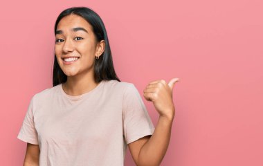 Asyalı genç bir kadın, günlük kıyafetler giyip gülümsüyor, gülümsüyor ve başparmağıyla yan tarafa bakıyor.. 