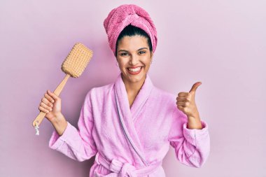 Duşta bornoz giyen, elinde sünger tutan, mutlu ve pozitif gülümseyen, başparmağıyla mükemmel ve onay işareti yapan genç İspanyol kadın. 