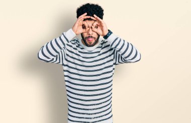 Sakallı, sıradan çizgili süveter giyen genç bir Arap dürbün gibi jest yapıyor, gözleri parmaklarından bakıyor. çılgın ifade. 