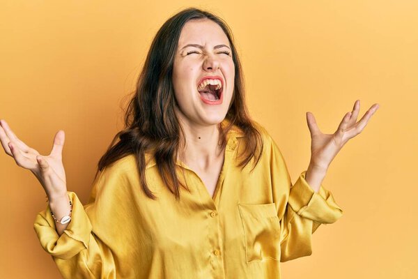 Young brunette woman wearing casual yellow shirt crazy and mad shouting and yelling with aggressive expression and arms raised. frustration concept. 