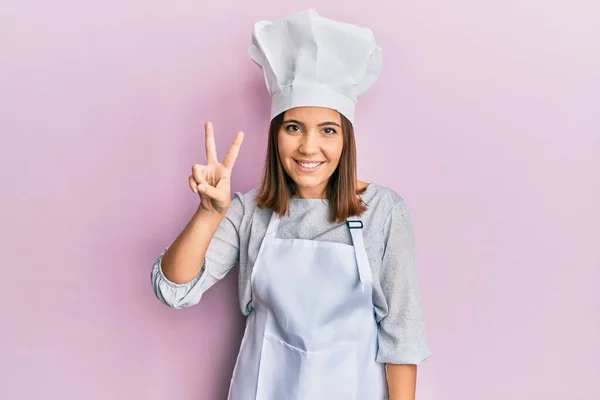 年轻而漂亮的女人穿着职业厨师的制服 头戴帽子 手指头一指二指 面带微笑 自信而快乐 — 图库照片