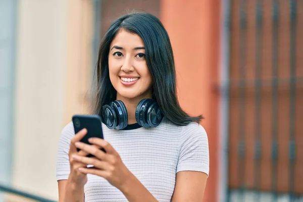 街中でスマホやヘッドフォンを使って笑顔で幸せそうな若いヒスパニック系女性 — ストック写真