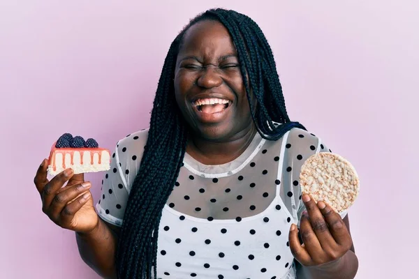 Young black woman with braids eating cake slice and rice crackers smiling and laughing hard out loud because funny crazy joke.