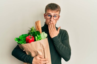 Kafkasyalı genç bir adam elinde torbayla market poşetleriyle ağzını eliyle kapatıyor, şokta ve hata yapmaktan korkuyor. şaşırmış ifade 