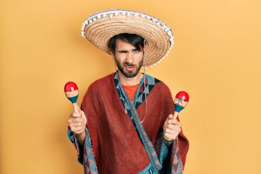 Meksikalı şapkalı genç İspanyol adam, elinde şüpheli ve gergin bir marakas tutuyor, sorun yüzünden surat asıyor. negatif kişi. 