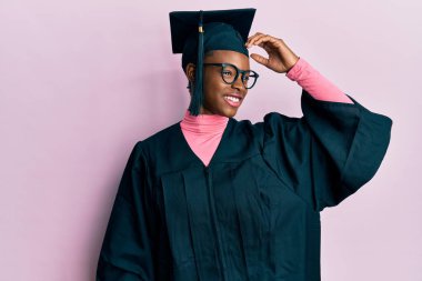 Mezuniyet şapkası ve tören cübbesi takan genç Afrikalı Amerikalı kız gülümseyerek saçlarına dokunarak el hareketleriyle çekici ve şık bir poz veriyor. 