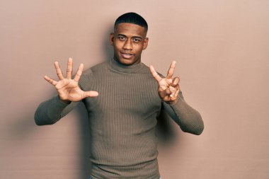 Balıkçı yaka kazak giyen genç siyahi bir adam yedi numaralı parmaklarını gösterip gösterirken kendine güvenen ve mutlu bir şekilde gülümsüyor.. 