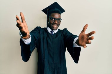 Mezuniyet şapkası ve tören cübbesi giymiş yakışıklı siyah adam kameraya sarılıp gülümsüyor. Mutluluğu kucaklayan neşeli bir ifade.. 