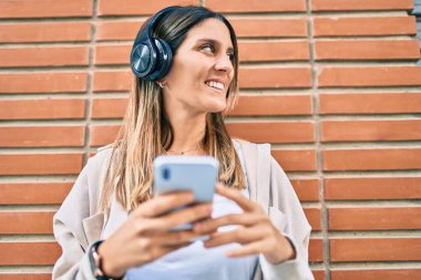 Beyaz kadın şehirde akıllı telefon ve kulaklık kullanarak mutlu bir şekilde gülümsüyor..