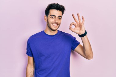 Gündelik tişört giyen genç İspanyol adam olumlu gülümsüyor ve el ve parmaklarıyla imza atıyor. başarılı ifade. 