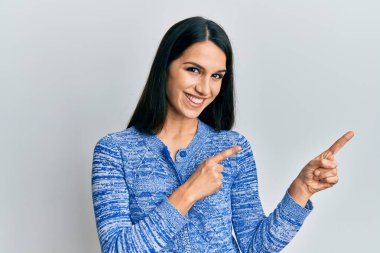 Gündelik kıyafetler giyen, gülümseyen ve kameraya bakan genç İspanyol kadın iki eliyle ve parmaklarıyla kenarı işaret ediyor.. 