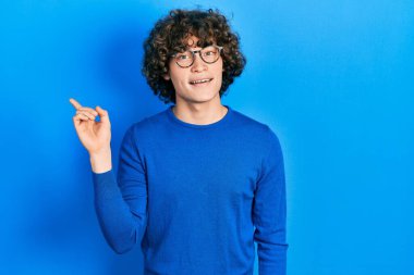 Yakışıklı, sıradan kıyafetler ve gözlük takan, yüzünde kocaman bir gülümseme olan, eli ve parmağı kameraya bakan genç bir adam.. 