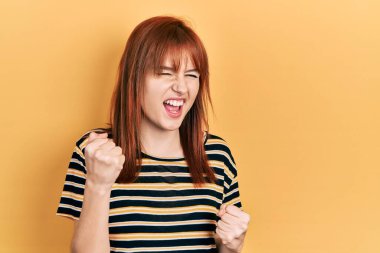 Kızıl saçlı genç kadın, sıradan çizgili tişört giyiyor. Şaşırmış ve şaşırmış bir şekilde kolları havada ve gözleri kapalı bir şekilde başarıyı kutluyor. 